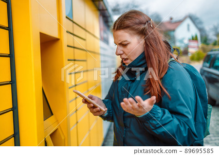 Caucasian angry woman with smartphone app at outdoor automated parcel machine. Mail delivery and post service concept Caucasian angry woman with smartphone app at outdoor automated parcel machine. Mail delivery and post service concept 89695585