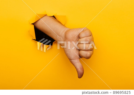 Hand showing a thumb down through ripped hole in yellow paper background. Concept of dislike and disapproval gesture 89695825