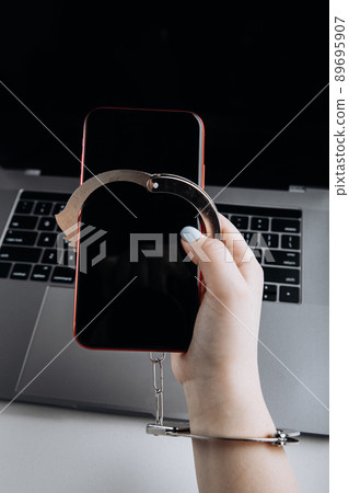 Concept of smartphone addiction. Phone chained to handcuff in hand with laptop background. Vertical image Concept of smartphone addiction. Phone chained to handcuff in hand with laptop background. Vertical image 89695907