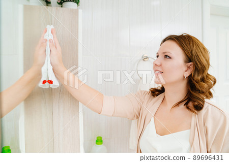 an attractive housewife washes the bathroom mirror with a special glass cleaner. an attractive housewife washes the bathroom mirror with a special glass cleaner. 89696431
