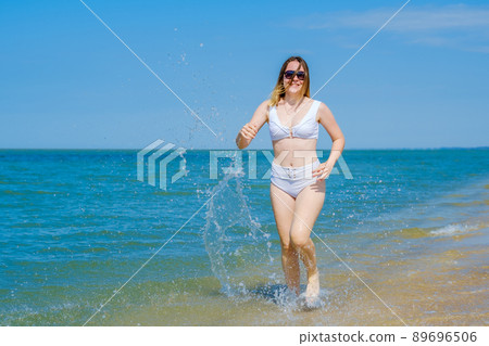 A young girl runs along the sandy sea beach along the surf. Splashes of sea water. The girl is wearing a white swimsuit and sunglasses. Freedom and carelessness. Outdoor fitness. Summer sunny day 89696506