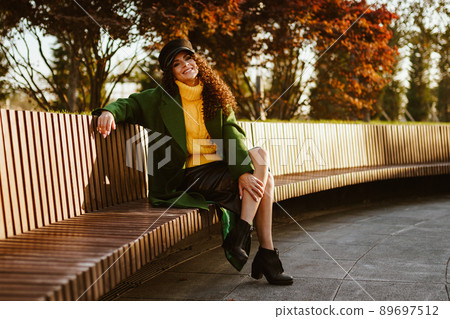 In the park on the bench sits in an autumn coat and cap a beautiful curly brunette with a charming smile 89697512