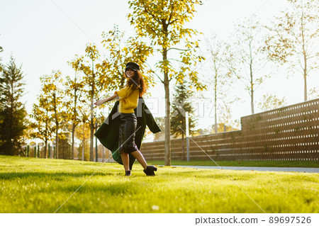 In the park on the green grass comes a girl in a bright sweater throwing on one shoulder coat In the park on the green grass comes a girl in a bright sweater throwing on one shoulder coat 89697526