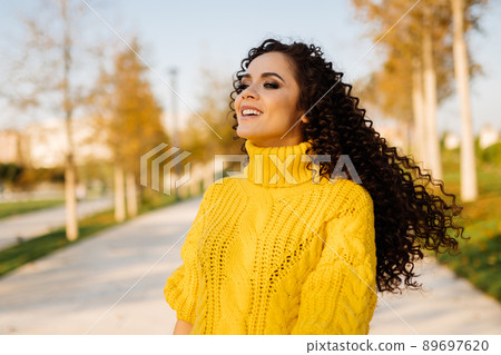 On the walkway in the park in a bright yellow sweater stands with fluttering hair a beautiful smiling girl 89697620