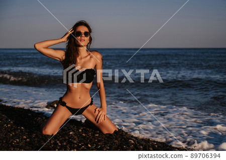 young girl with a sexy body in black swimsuit and sunglasses erotically sits on knees on beach by sea young girl with a sexy body in black swimsuit and sunglasses erotically sits on knees on beach by sea 89706394