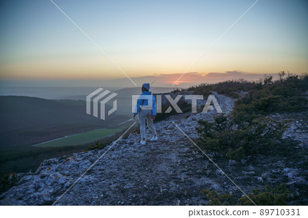 Woman tourist on top of sunrise mountain. The girl salutes the sun, wearing a blue jacket, white hat and white jeans. Conceptual design. 89710331