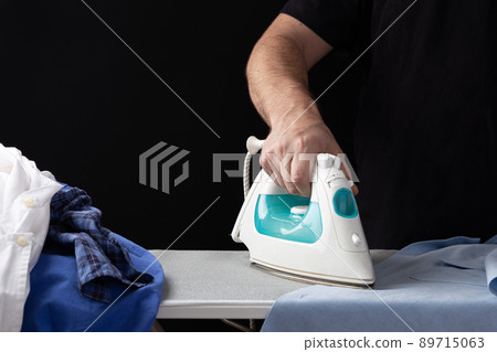 A man's hand with an iron, ironing shirts on the ironing table, on a dark background. A man's hand with an iron, ironing shirts on the ironing table, on a dark background. 89715063