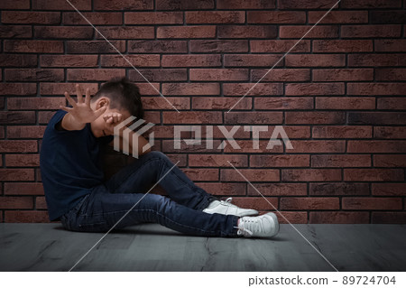 Scared little boy closing eyes with hand on floor near brick wall. Child in danger 89724704
