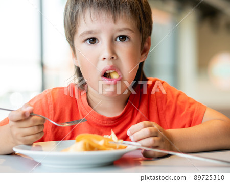 Kid in red t-shirt eats food with knife and fork. Fastidious boy with puzzled expression on face. 89725310
