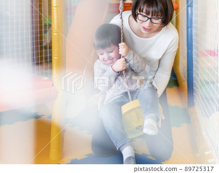 Toddler plays on rope swing with his mother or babysitter. Physical development for little children. Interior of kindergarten or nursery. Toddler plays on rope swing with his mother or babysitter. Physical development for little children. Interior of kindergarten or nursery. 89725317