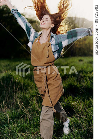 Woman walking on the grass in nature smiling in farm clothes with beautiful hair flying in the wind happy in the rays of summer sunset light Woman walking on the grass in nature smiling in farm clothes with beautiful hair flying in the wind happy in the rays of summer sunset light 89725982