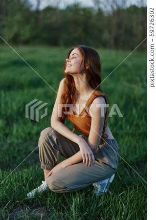 A beautiful redheaded woman sits on the grass in sporty summer clothes and relaxes after a walk in nature and looks out at the sunset. 89726302