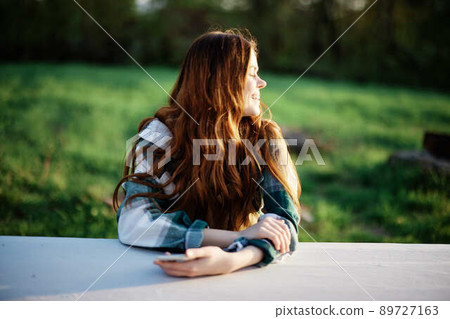 Girl with phone in hand sitting in nature in the park at the table smiling beautifully and looking at the camera with her red hair lit by the sunset sunlight of summer 89727163