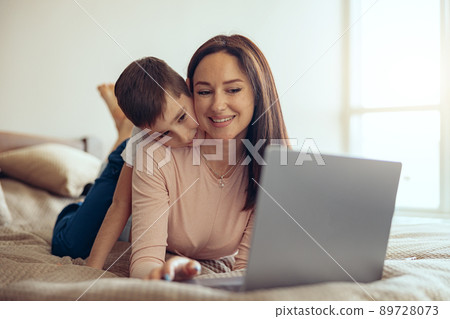 Happy beautiful young Caucasian woman lying on bed with laptop, small son hugging behind. 89728073