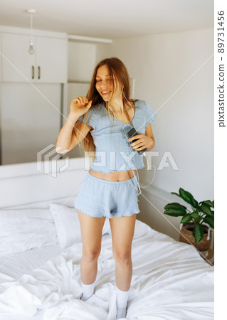 Young teen girl in pajama standing on her bed and singing a song. Cheerful caucasian adolescent girl with headphones dancing on her bedroom . Young teen girl in pajama standing on her bed and singing a song. Cheerful caucasian adolescent girl with headphones dancing on her bedroom . 89731456