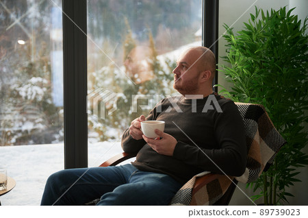 Young man enjoying weekends inside contemporary barn house. Male tourist sitting on chair, holding cup of tea, enjoying view of mountain landscape through panoramic windows in new cottage. Young man enjoying weekends inside contemporary barn house. Male tourist sitting on chair, holding cup of tea, enjoying view of mountain landscape through panoramic windows in new cottage. 89739023