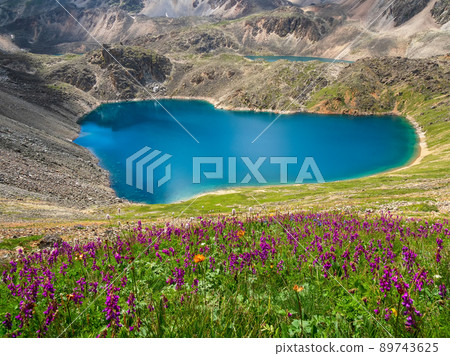 Bright summer azure mountain lake among black rocks. Blue alpine lake among sunlit black green rocky flower hills in highlands. 89743625