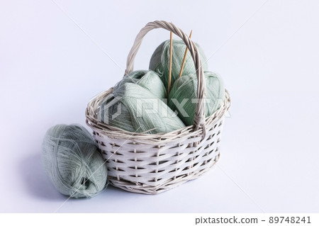 Light green yarn threads in a white basket on a blue background with knitting needles. Light green yarn threads in a white basket on a blue background with knitting needles. 89748241