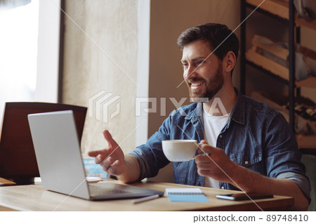 Handsome cheerful young man videochatting at laptop in cafe and drinking coffee. Video call. 89748410