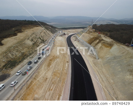 yellow truck pouring bitumen over a new road. Aerial drone shot. Road construction in progress on slope nature canyon. Infrastructure development and logistics. 89748429
