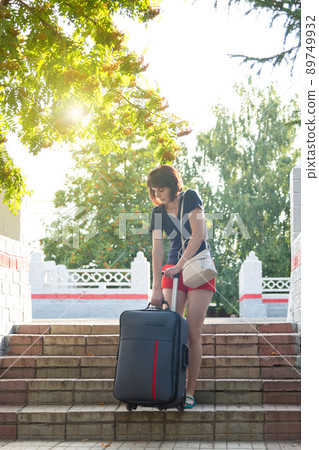 A brunette woman in red shorts hardly lifts a large heavy suitcase up the steps at the train station A brunette woman in red shorts hardly lifts a large heavy suitcase up the steps at the train station 89749932