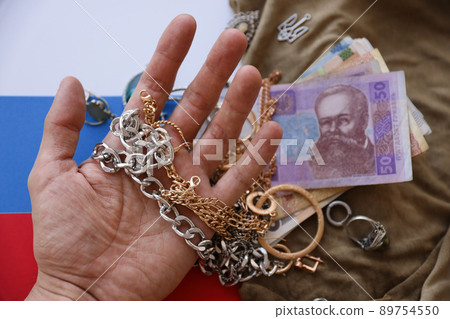 Marauders hand with bunch of stolen jewelry and money on military uniform cloth fabric and Russian flag. Looting by Russian soldiers in the Ukrainian cities Marauders hand with bunch of stolen jewelry and money on military uniform cloth fabric and Russian flag. Looting by Russian soldiers in the Ukrainian cities 89754550