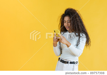Curly-haired young woman using her cell phone with yellow background Curly-haired young woman using her cell phone with yellow background 89757308