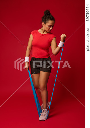 Young multi-ethnic sports fitness woman in sportswear working out isolated on red background in studio. Sport exercises healthy lifestyle concept. Doing exercises with elastic band. 89759634