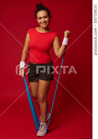 Attractive athletic fit woman doing body weight workout using elastic strap pulling with arms for shoulder training against red background with copy ad space 89759635