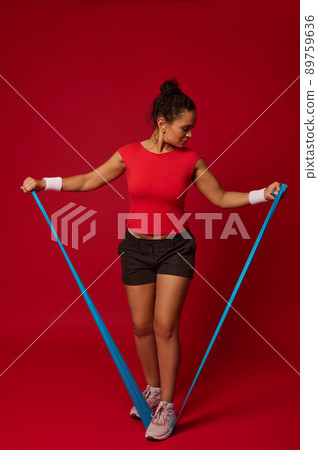 Full body size portrait of a sporty woman exercising with elastic fitness band while performing functional body weight training against res colored background with copy ad space Full body size portrait of a sporty woman exercising with elastic fitness band while performing functional body weight training against res colored background with copy ad space 89759636
