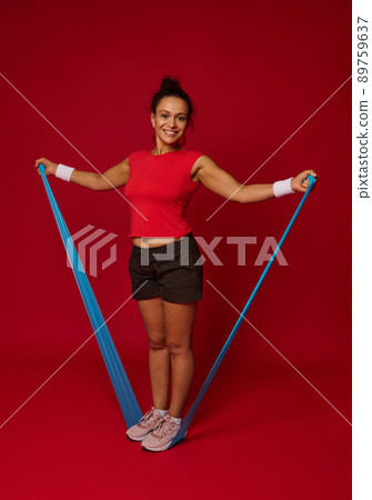 Full length of a sporty woman with exercise band against red background with copy space for advertisement text. Fitness, sport, active and healthy lifestyle concept Full length of a sporty woman with exercise band against red background with copy space for advertisement text. Fitness, sport, active and healthy lifestyle concept 89759637