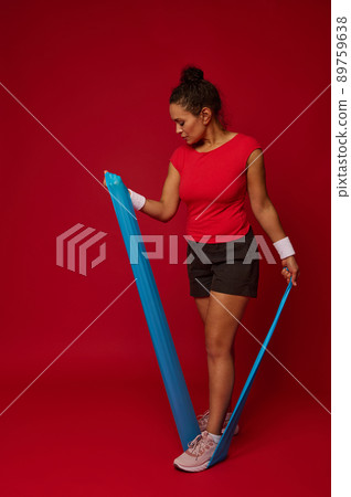 Full length of a sporty woman using elastic fitness rubber band exercises against red background with copy space for advertisement text. Fitness, sport, active and healthy lifestyle concept Full length of a sporty woman using elastic fitness rubber band exercises against red background with copy space for advertisement text. Fitness, sport, active and healthy lifestyle concept 89759638