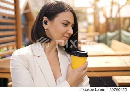 A thoughtful business woman in a white jacket with a disposable cup of tea looks away. A free moment to relax. Working Coffee Break. The person listens to music using wireless earphones A thoughtful business woman in a white jacket with a disposable cup of tea looks away. A free moment to relax. Working Coffee Break. The person listens to music using wireless earphones 89760455