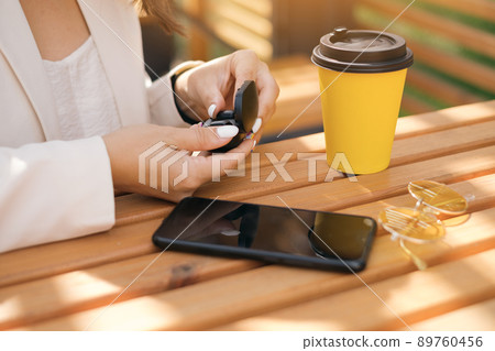 Close-up of a women's hands taking out of the box wireless small black earbuds. Girl at a table with a black mobile smartphone and a glass of coffee and sunglasses . White manicure 89760456