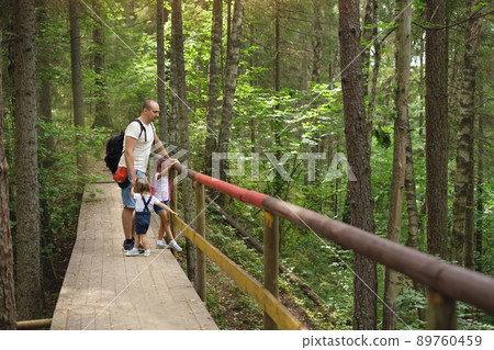 Dad and daughters are standing on old wooden footbridge in woods among trees. Hiking with whole family with small kid. Traveling through ecological trail. Man with hiking backpack holds girls by hands Dad and daughters are standing on old wooden footbridge in woods among trees. Hiking with whole family with small kid. Traveling through ecological trail. Man with hiking backpack holds girls by hands 89760459