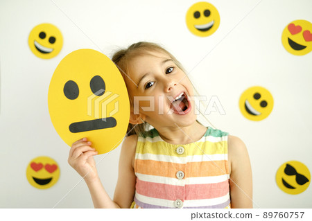 The girl holds a sad emoticon face in hand, but she smiling and laughs no matter what. A cheerful happy sincere child The girl holds a sad emoticon face in hand, but she smiling and laughs no matter what. A cheerful happy sincere child 89760757