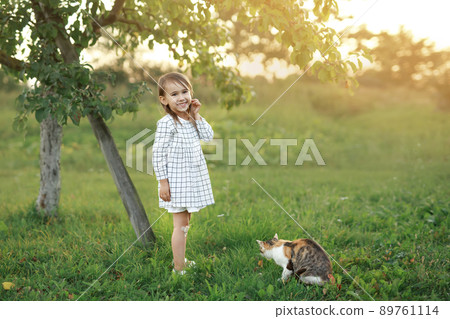 A child in the garden stands smiling sweetly next to a stray cat at sunset. Pets and their owners. Friendship of children and animals. A child in the garden stands smiling sweetly next to a stray cat at sunset. Pets and their owners. Friendship of children and animals. 89761114