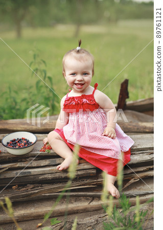 A little one-year-old girl in a red dress is sitting in the garden on old boards and eating currant and blueberry berries. The season of wild berries. Summer warm laziness in nature in the fresh air 89761121