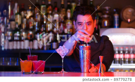 Young bartender mixing ingredients in the shaker with a spoon - a glass with ice - cocktails on the stand 89770278