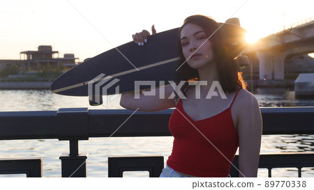 Young attractive woman standing on the waterfront on sunset holding a skateboard - posing on the camera Young attractive woman standing on the waterfront on sunset holding a skateboard - posing on the camera 89770338
