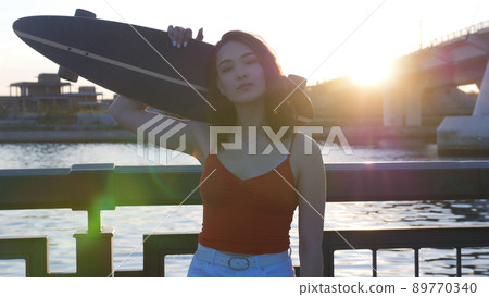 Young woman standing on the waterfront on sunset holding a skateboard - posing on the camera Young woman standing on the waterfront on sunset holding a skateboard - posing on the camera 89770340