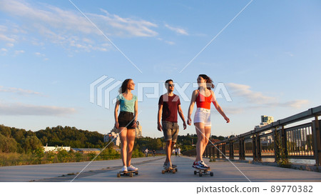 Young friends riding skateboards on the waterfront on a background of modern buildings - sunset Young friends riding skateboards on the waterfront on a background of modern buildings - sunset 89770382