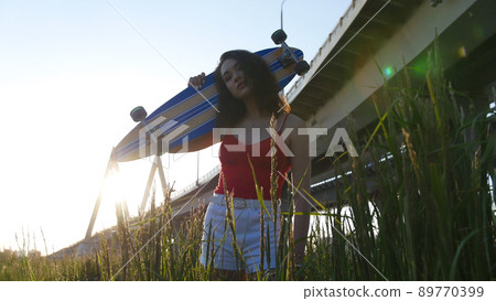 Young smiling woman standing in the high grass and holding a skateboard Young smiling woman standing in the high grass and holding a skateboard 89770399