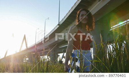 Young smiling woman with skateboard standing in the high grass and posing for the camera Young smiling woman with skateboard standing in the high grass and posing for the camera 89770401