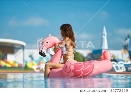 Dreamy slim female sitting on float in pool with clean blue water at resort. Side view of long haired girl floating on pink inflatable flamingo in pool on sunny summer day. Concept of entertainment. Dreamy slim female sitting on float in pool with clean blue water at resort. Side view of long haired girl floating on pink inflatable flamingo in pool on sunny summer day. Concept of entertainment. 89772329
