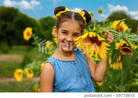 A happy little girl in a blue dress stands next to the flowers of a decorative sunflower 89778658