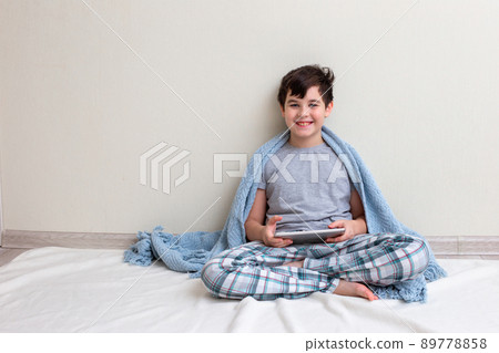 A handsome smiling boy, a teenager in gray pajamas, in a blue knitted plaid sits on the floor, holds a digital tablet A handsome smiling boy, a teenager in gray pajamas, in a blue knitted plaid sits on the floor, holds a digital tablet 89778858