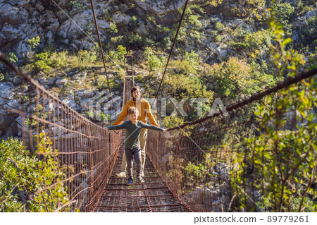 Father and son tourists on Old rusty bridge. Attraction Long extreme suspension iron bridge across the river Moraca. Sights of Montenegro. Landmark Montenegro Father and son tourists on Old rusty bridge. Attraction Long extreme suspension iron bridge across the river Moraca. Sights of Montenegro. Landmark Montenegro 89779261