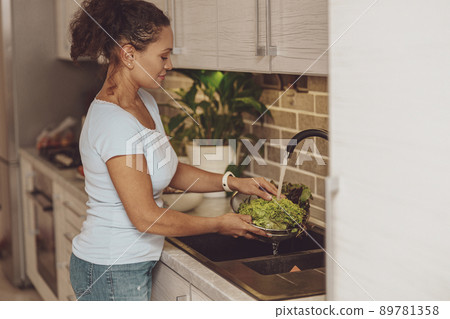 A woman standing in front of the sink in the kitchen washes greens A woman standing in front of the sink in the kitchen washes greens 89781358