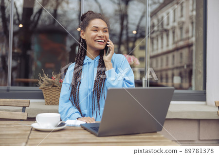 Cheerful young mixed-race stylish woman chatting on phone with coffee while working on laptop. 89781378
